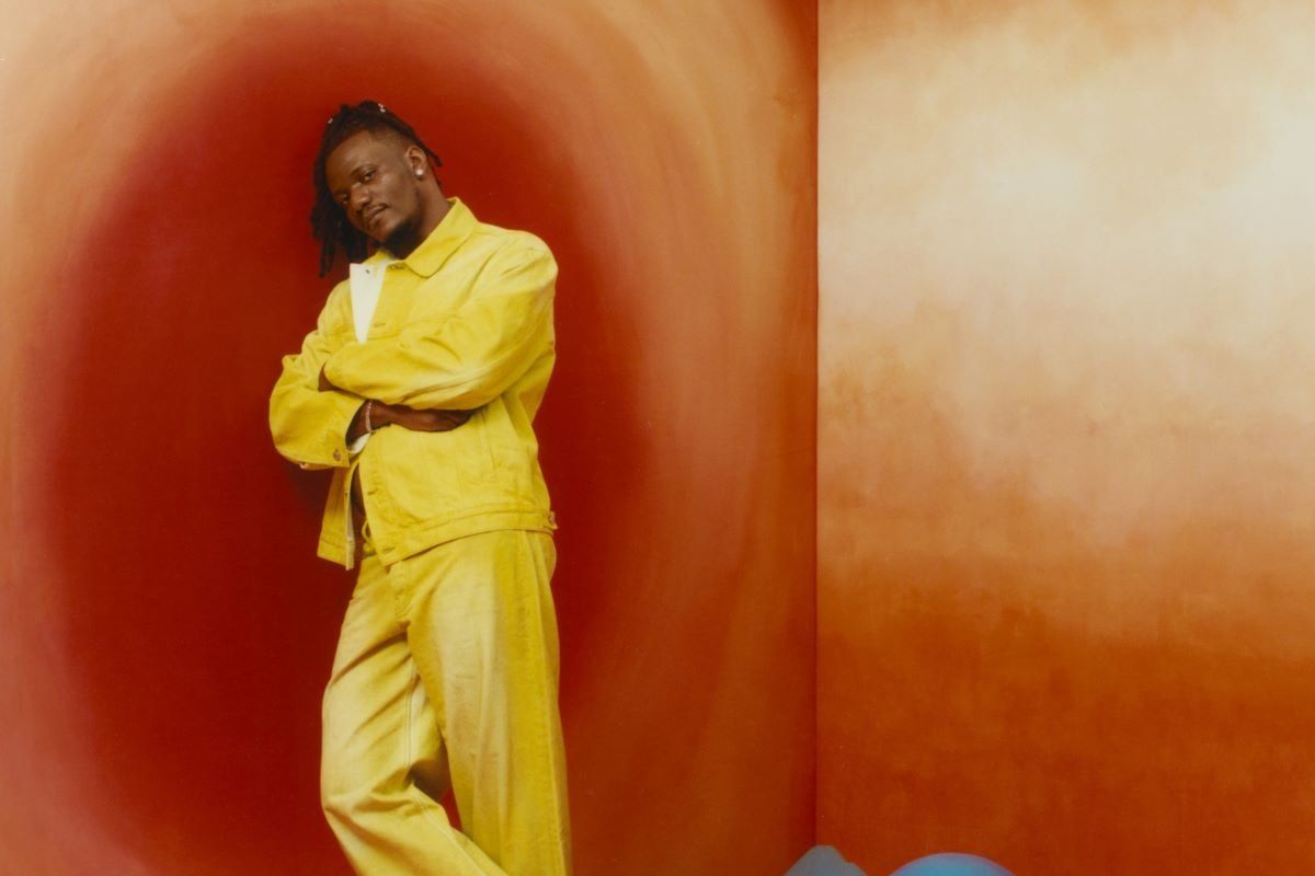 A photo of Pheelz resting his shoulders on an orange-coloured wall with arms crossed.